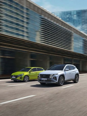 Hyundai i20 in Gelb und Hyundai TUCSON in Silber fahren vor moderner Stadtkulisse. Promotionbild für günstige Leasingangebote beider Hyundai Modelle.