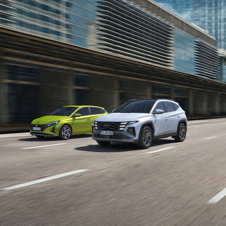 Hyundai i20 in Gelb und Hyundai TUCSON in Silber fahren vor moderner Stadtkulisse. Promotionbild für Leasingangebote des Hyundai i20 und Hyundai TUCSON zu attraktiven Konditionen.